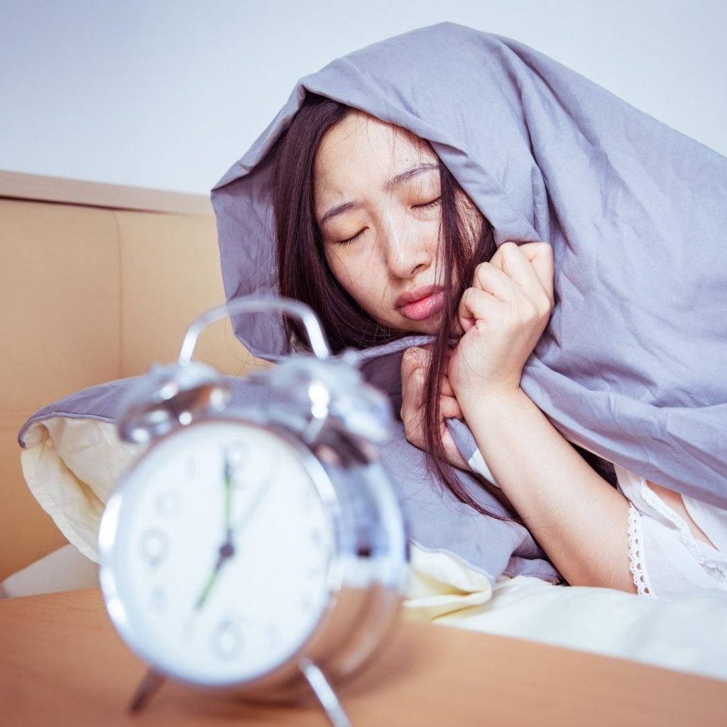 ADHD Life Hacks for better mornings: A woman is struggling to wake up, lying in bed with an alarm clock in the foreground. 